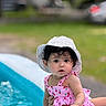 Aitana is registered to the contest to win money with this photo: baby, child, swimsuit, pink, hat, white_hat, curly_hair, pool, water, outdoor, summer, cute, portrait, sitting, person, infant, green_background, daylight, skin, face