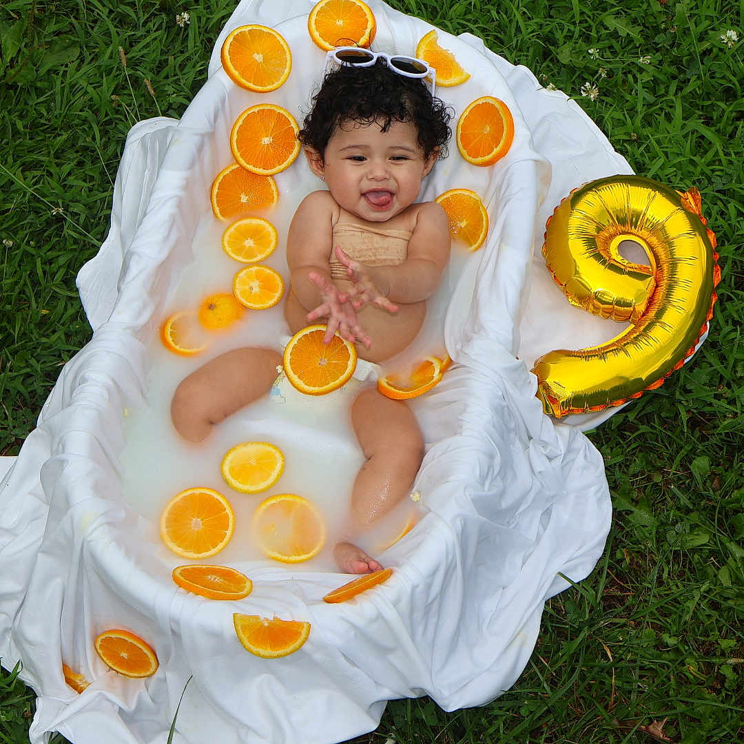 Aitana is registered to the contest to win money with this photo: baby, bathtub, orange_slices, milky_water, grass, balloon, number_9, sunglasses, curly_hair, smiling, outdoor, white_cloth, playful, child, summer, cute, sitting, fun, portrait, nature