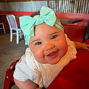 Everly is registered to the contest to win money with this photo: baby, child, smile, bow, headband, high_chair, restaurant, table, red, white_clothing, happy, face, cute, infant, curly_hair, interior, wooden_chair, person, closeup, portrait