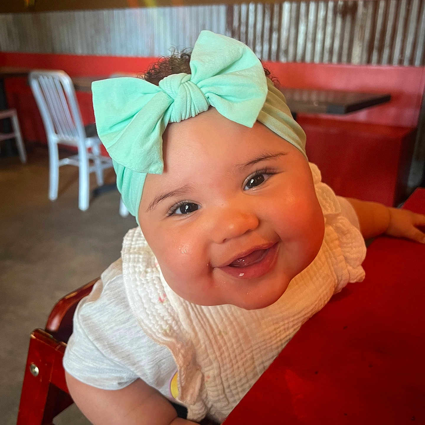 Everly is registered to the contest to win money with this photo: baby, child, smile, bow, headband, high_chair, restaurant, table, red, white_clothing, happy, face, cute, infant, curly_hair, interior, wooden_chair, person, closeup, portrait