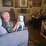 man, dog, couch, living_room, wall_art, framed_picture, clock, decorations, furniture, carpet, seated, petting, indoor, casual_clothing, home, relaxing, white_dog, older_adult, calm, comfortable