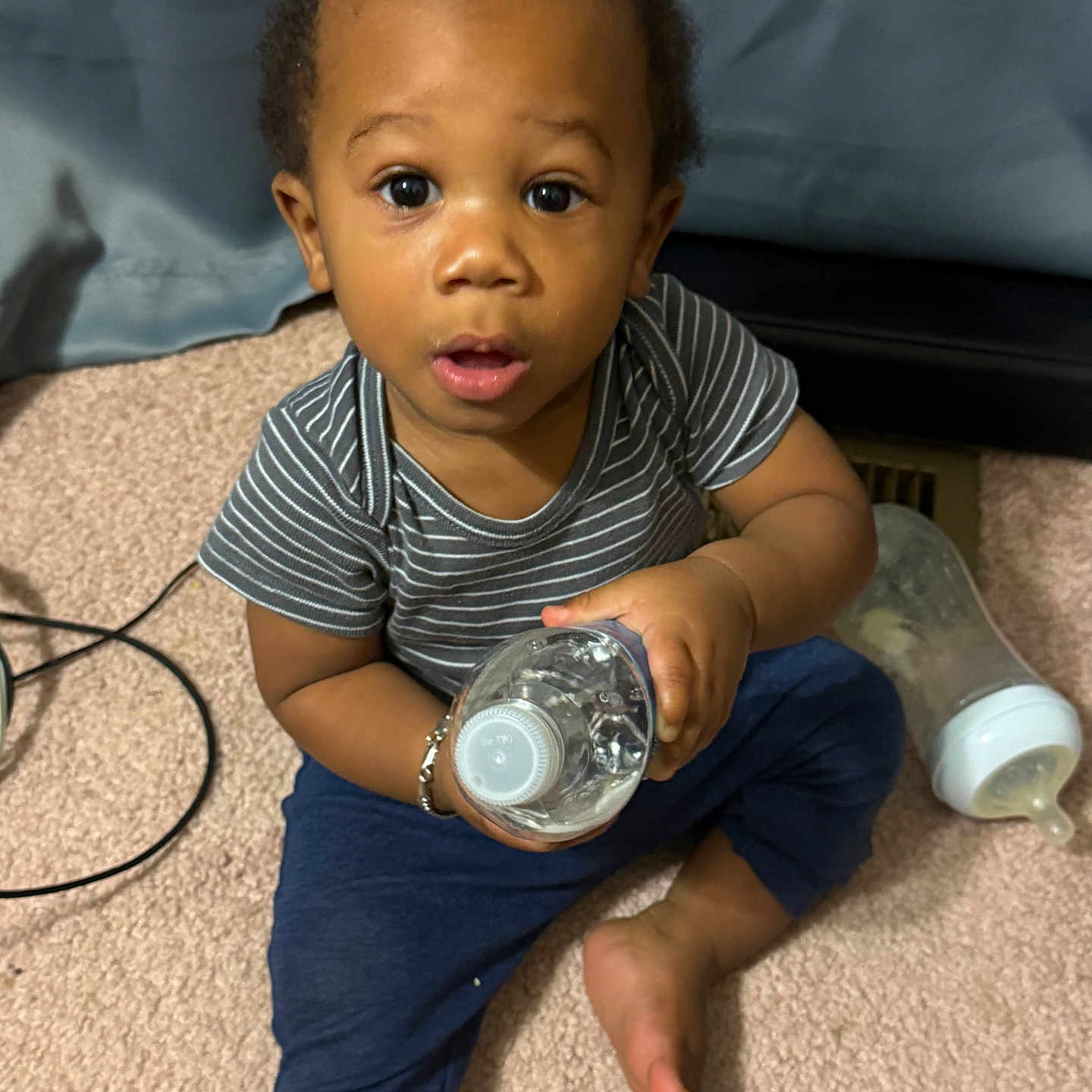Clayton joined the competition — help win amazing prizes! baby, child, toddler, sitting, carpet, indoors, portrait, face, wide_eyed, curious, water_bottle, baby_bottle, striped_shirt, blue_pants, barefoot, bedroom, furniture, floor, candid, home