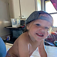 Joaleen is registered to the contest to win money with this photo: toddler, child, smile, cap, baseball_cap, indoor, kitchen, window, natural_light, happy, person, face, skin, hand, table, diaper, curtain, cabinet, mug, container