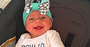 Andi is registered to the contest to win money with this photo: baby, infant, child, smiling, happy, portrait, car_seat, vehicle, bow, hat, baby_clothing, white_shirt, text_on_clothing, sitting, face, eyes, mouth, indoor, close_up, safe