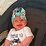 baby, infant, child, smiling, happy, portrait, car_seat, vehicle, bow, hat, baby_clothing, white_shirt, text_on_clothing, sitting, face, eyes, mouth, indoor, close_up, safe