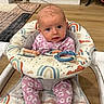 Andi is registered to the contest to win money with this photo: baby, infant, child, sitting, activity_walker, toy, colorful, pink_clothing, patterned_clothing, face, portrait, indoors, living_room, bookshelf, books, wooden_floor, playtime, curious, household, rainbow_pattern