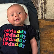 Andi is registered to the contest to win money with this photo: baby, infant, child, portrait, smiling, lying_down, changing_pad, black_shirt, text_on_clothing, daddy, colorful_letters, wooden_floor, patterned_rug, indoor, face, eyes, mouth, happy, casual, photography