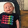 baby, infant, child, portrait, smiling, lying_down, changing_pad, black_shirt, text_on_clothing, daddy, colorful_letters, wooden_floor, patterned_rug, indoor, face, eyes, mouth, happy, casual, photography