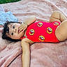 Daniela is registered to the contest to win money with this photo: baby, child, infant, swimsuit, red_clothing, bracelet, blanket, pink_blanket, grass, outdoor, playing, lying_down, cute, curly_hair, footwear, clothes, hand_near_mouth, portrait, young_child, relaxing