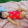 baby, child, curly_hair, swimsuit, red_clothing, blanket, outdoor, grass, laying_down, infant, cute, portrait, relaxing, summer, skin, person, young_child, playful, casual, resting