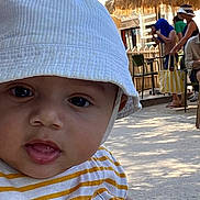 Gabriel a rejoint le concours — aidez-le/la à gagner de superbes lots ! baby, child, face, hat, bucket_hat, striped_shirt, yellow, white, outdoor, daylight, people, background, blur, seated, casual, expression, curious, portrait, closeup, shade