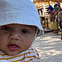 baby, child, face, hat, bucket_hat, striped_shirt, yellow, white, outdoor, daylight, people, background, blur, seated, casual, expression, curious, portrait, closeup, shade