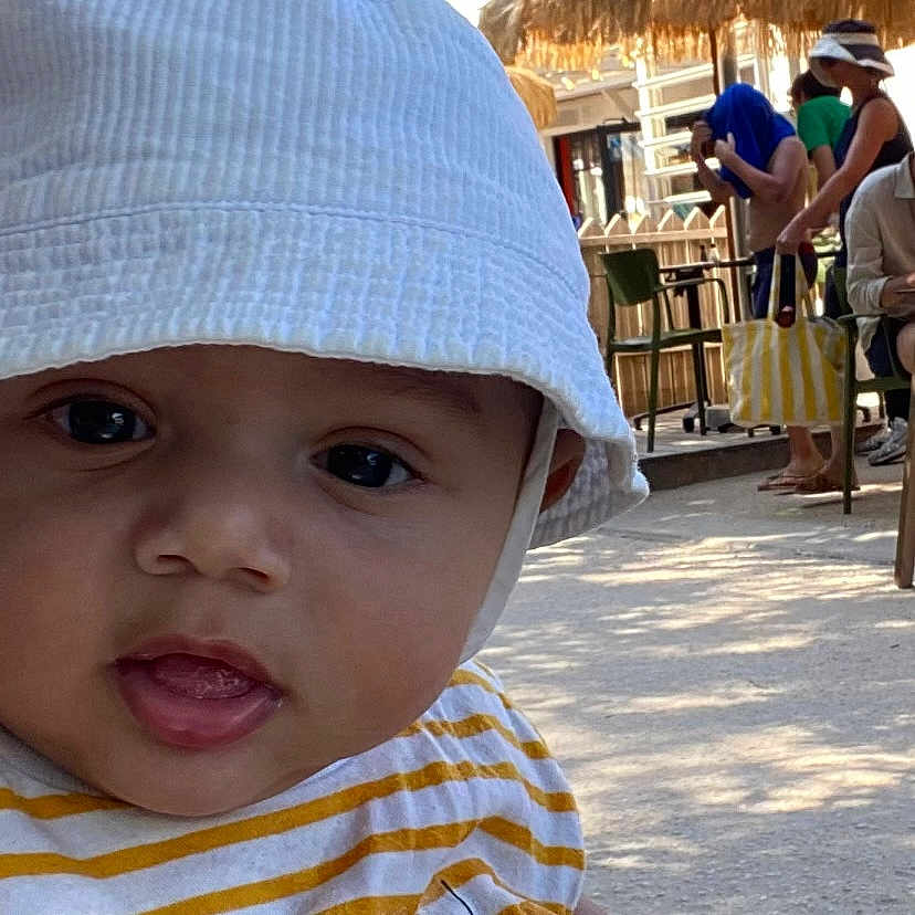 Gabriel a rejoint le concours — aidez-le/la à gagner de superbes lots ! baby, child, face, hat, bucket_hat, striped_shirt, yellow, white, outdoor, daylight, people, background, blur, seated, casual, expression, curious, portrait, closeup, shade