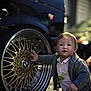 toddler, child, car_wheel, bmw, golden_wheel, sweater, outdoor, night, curious, baby, wheel_rim, automobile, hand, young_child, clothing, portrait, light, reflection, street, closeup