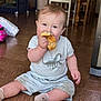 baby, child, indoor, floor, tile_floor, eating, croissant, dinosaur_outfit, socks, seated, smiling, cute, messy, furniture, table, chair, toy, home, happy, portrait