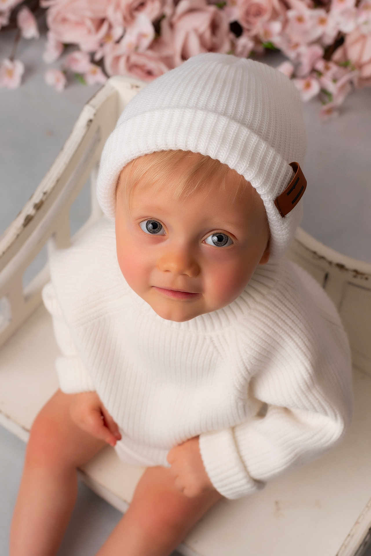 Camille participe au concours pour gagner de l'argent avec cette photo : toddler, child, white_hat, white_sweater, beanie, bench, pink_flowers, portrait, indoor, cute, soft_lighting, smiling, baby, face, looking_up, hands, skin, cozy, warm, innocent