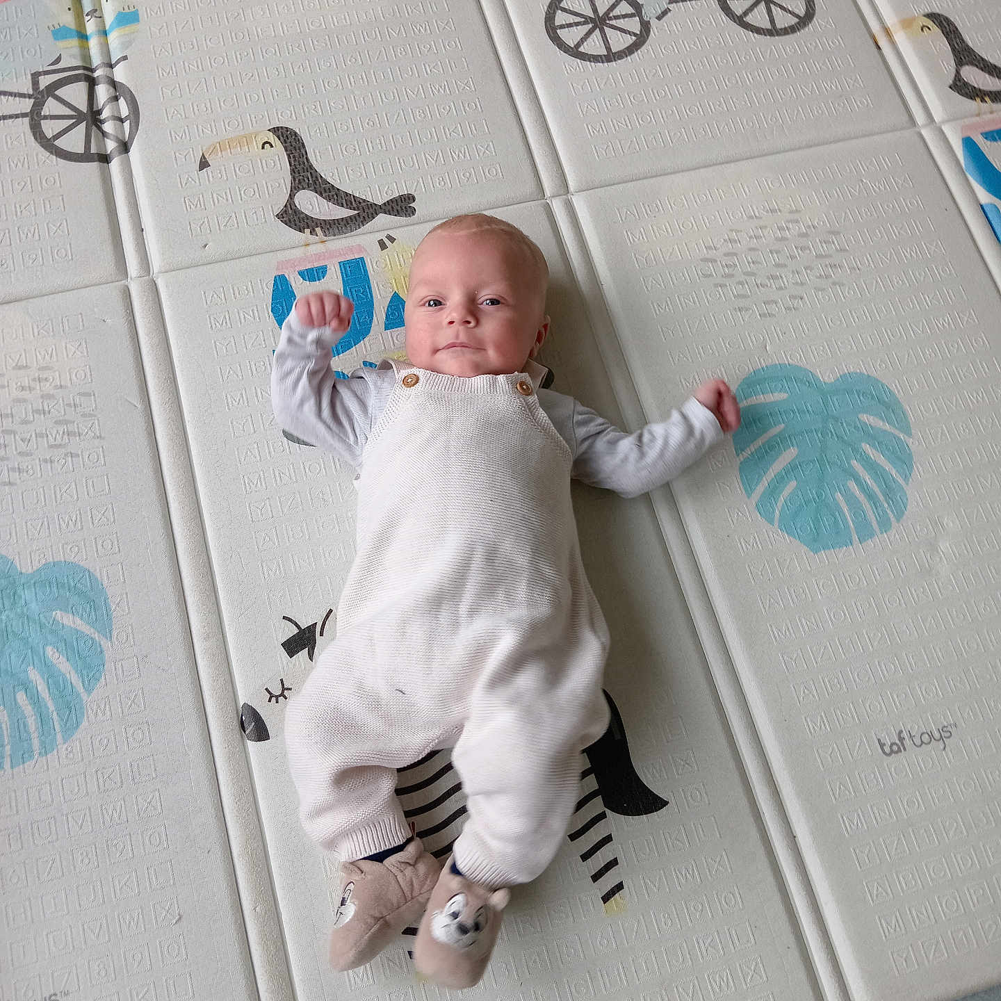 Aaron participe au concours pour gagner de l'argent avec cette photo : baby, infant, play_mat, floor, white_overalls, slippers, animal_pattern, leaf_pattern, cute, child, indoors, smiling, lying_down, soft_lighting, playful, young_child, person, baby_clothes, floor_mat, cozy