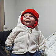 Aaron participe au concours pour gagner de l'argent avec cette photo : baby, child, red_hat, white_jacket, gloves, blue_pants, indoor, smiling, happy, warm_clothing, cozy, seated, person, infant, cute, portrait, headwear, winter_clothes, home, blanket