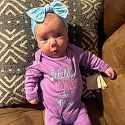 Mia is registered to the contest to win money with this photo: baby, infant, purple_onesie, headband, blue_bow, cushion, patterned_cushion, couch, brown_couch, face, person, cute, expression, indoor, child, newborn, clothing, portrait, lying_down, looking_at_camera