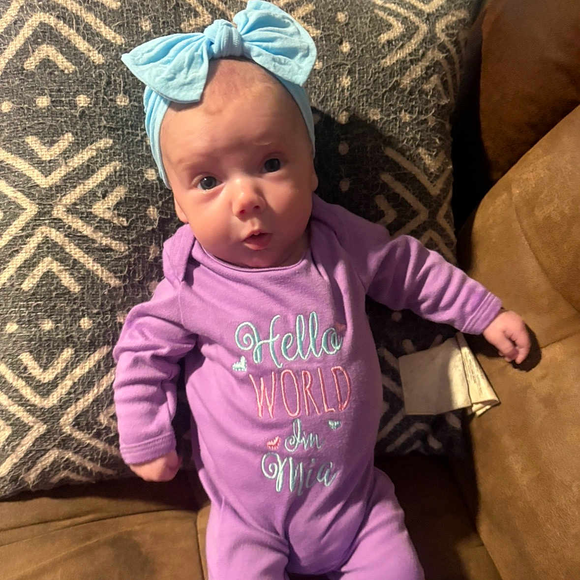 Mia is registered to the contest to win money with this photo: baby, infant, purple_onesie, headband, blue_bow, cushion, patterned_cushion, couch, brown_couch, face, person, cute, expression, indoor, child, newborn, clothing, portrait, lying_down, looking_at_camera