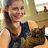 woman, cat, pet, portrait, smiling, indoors, cuddling, sitting, brown_cat, long_hair, ponytail, black_shirt, arms, living_room, window, warm_lighting, furniture, happy, animal, human