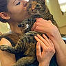 woman, person, cat, tabby_cat, pet, animal, portrait, indoors, hugging, kissing, affection, whiskers, green_eyes, striped_fur, hands, arms, long_hair, earrings, warm_lighting, close_up