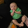 baby, infant, child, green_shirt, black_shorts, couch, blanket, hand_in_mouth, indoors, furniture, person, sitting, cute, young, skin, face, foot, relaxing, home, casual