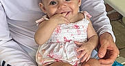 Zavia is registered to the contest to win money with this photo: baby, child, smiling, person, adult, holding, dress, sitting, bench, brick_wall, hand, finger_in_mouth, white_shirt, blue_shorts, patterned_clothing, happy, outdoor, skin, face, cute