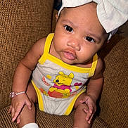 Sienna is registered to the contest to win money with this photo: baby, child, infant, onesie, headband, bow, winnie_the_pooh, sitting, brown_chair, white_socks, cute, portrait, indoors, person, young_child, expression, looking_at_camera, skin, hand, foot