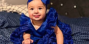 Mia is registered to the contest to win money with this photo: baby, child, blue_dress, headband, bow, bedspread, pattern, happy, smiling, tongue_out, sitting, indoor, cute, infant, hands, feet, soft_focus, bed, fabric, portrait