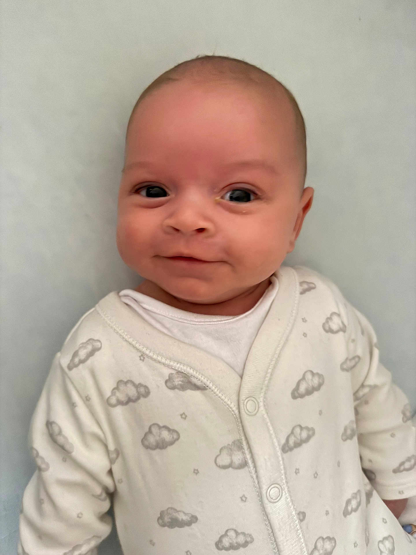 Theo is registered to the contest to win money with this photo: baby, infant, smiling, face, eyes, onesie, cloud_pattern, white_clothing, cute, portrait, child, newborn, soft_background, lying_down, small_hands, cheeks, happy, indoors, person, closeup