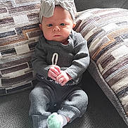 Gabriella is registered to the contest to win money with this photo: baby, infant, child, headband, bow, grey_outfit, couch, cushion, patterned_fabric, hands, sitting, indoors, carpet, portrait, face, expression, feet, sock, cozy, furniture
