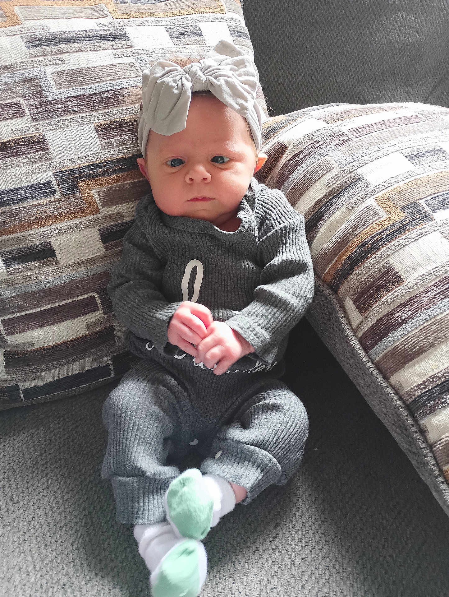 Gabriella is registered to the contest to win money with this photo: baby, infant, child, headband, bow, grey_outfit, couch, cushion, patterned_fabric, hands, sitting, indoors, carpet, portrait, face, expression, feet, sock, cozy, furniture