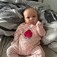 Olivia is registered to the contest to win money with this photo: baby, infant, pink_onesie, hearts, bed, blanket, patterned_blanket, tongue_out, cute, child, indoors, person, sleepwear, soft_lighting, home, playful, lying_down, newborn, small_person, comfort