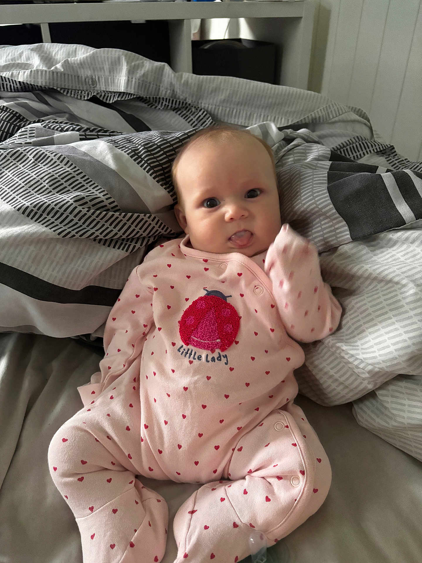 Olivia is registered to the contest to win money with this photo: baby, infant, pink_onesie, hearts, bed, blanket, patterned_blanket, tongue_out, cute, child, indoors, person, sleepwear, soft_lighting, home, playful, lying_down, newborn, small_person, comfort