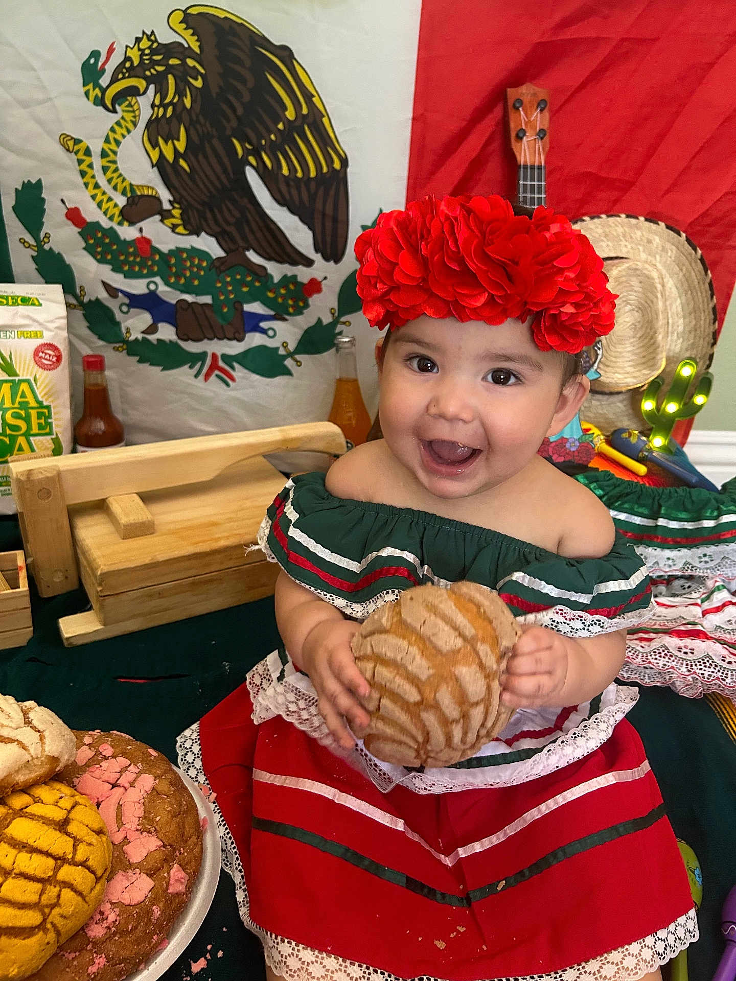 Hailey joined the competition — help win amazing prizes! bread, celebration, child, concha_bread, decorations, food, green, guitar, happy, indoors, mexican_flag, pastry, red, red_flower_headband, smiling, sombrero, table, toddler, traditional_clothing, white
