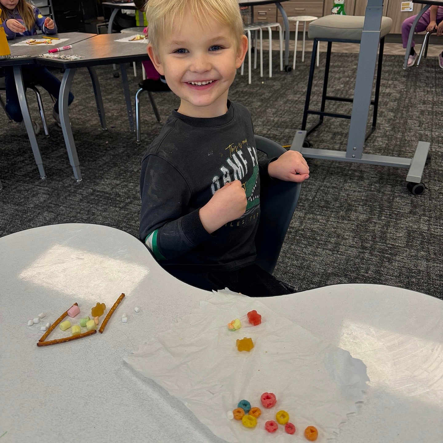 Dax is registered to the contest to win money with this photo: child, boy, classroom, table, snacks, pretzel, cereal, napkin, smile, happy, indoor, education, learning, kids, furniture, carpet, chair, clothing, person, people