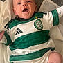 baby, infant, child, smiling, green, white, clothing, sportswear, football_jersey, celtic_football_club, socks, blanket, soft_surface, happy, lying_down, cute, portrait, indoor, face, person