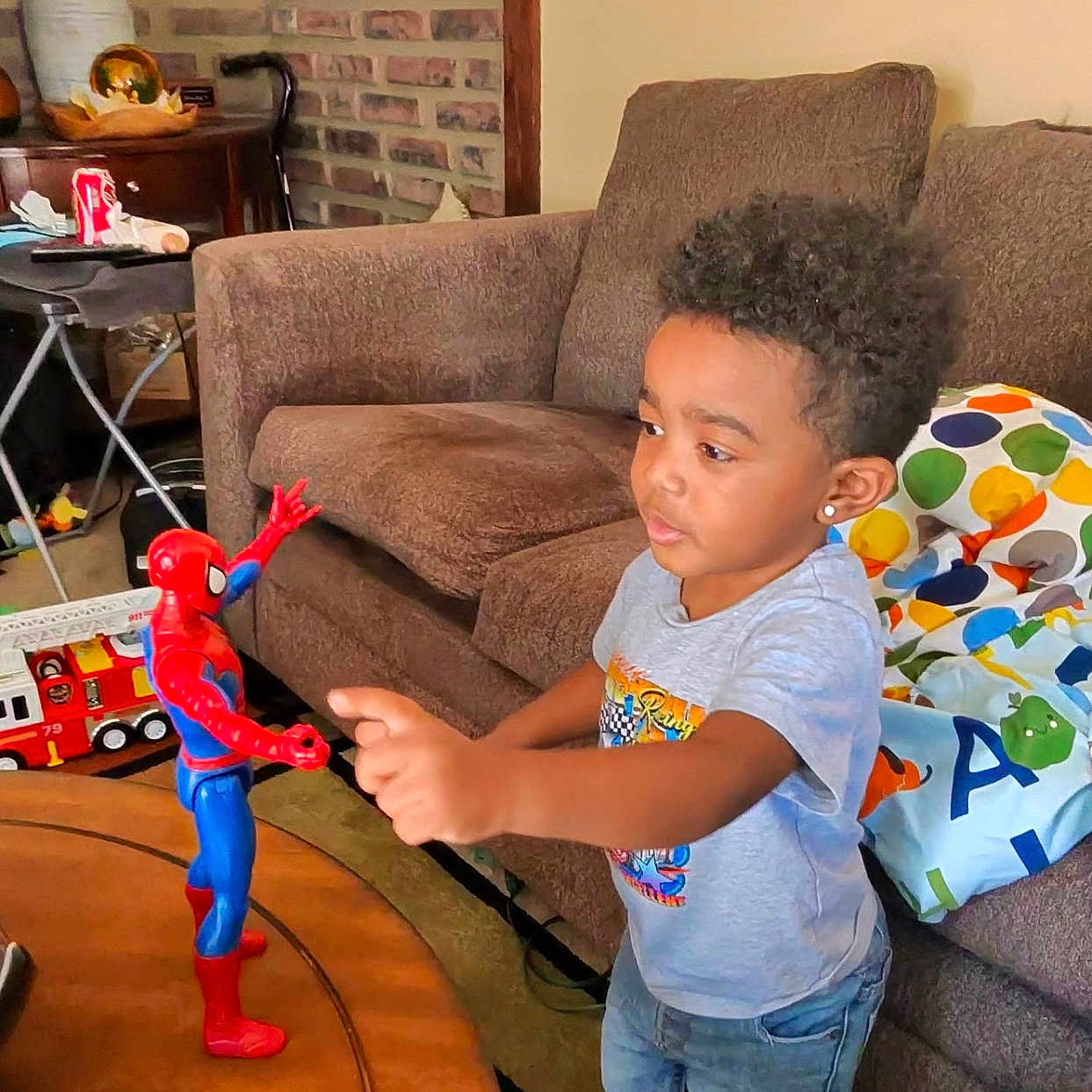 Zander is registered to the contest to win money with this photo: action_figure, barefoot, boy, brick_wall, casual_clothing, child, couch, curly_hair, diaper_pack, furniture, home, indoor, interaction, lamp, living_room, pillow, play, spider_man, table, toy