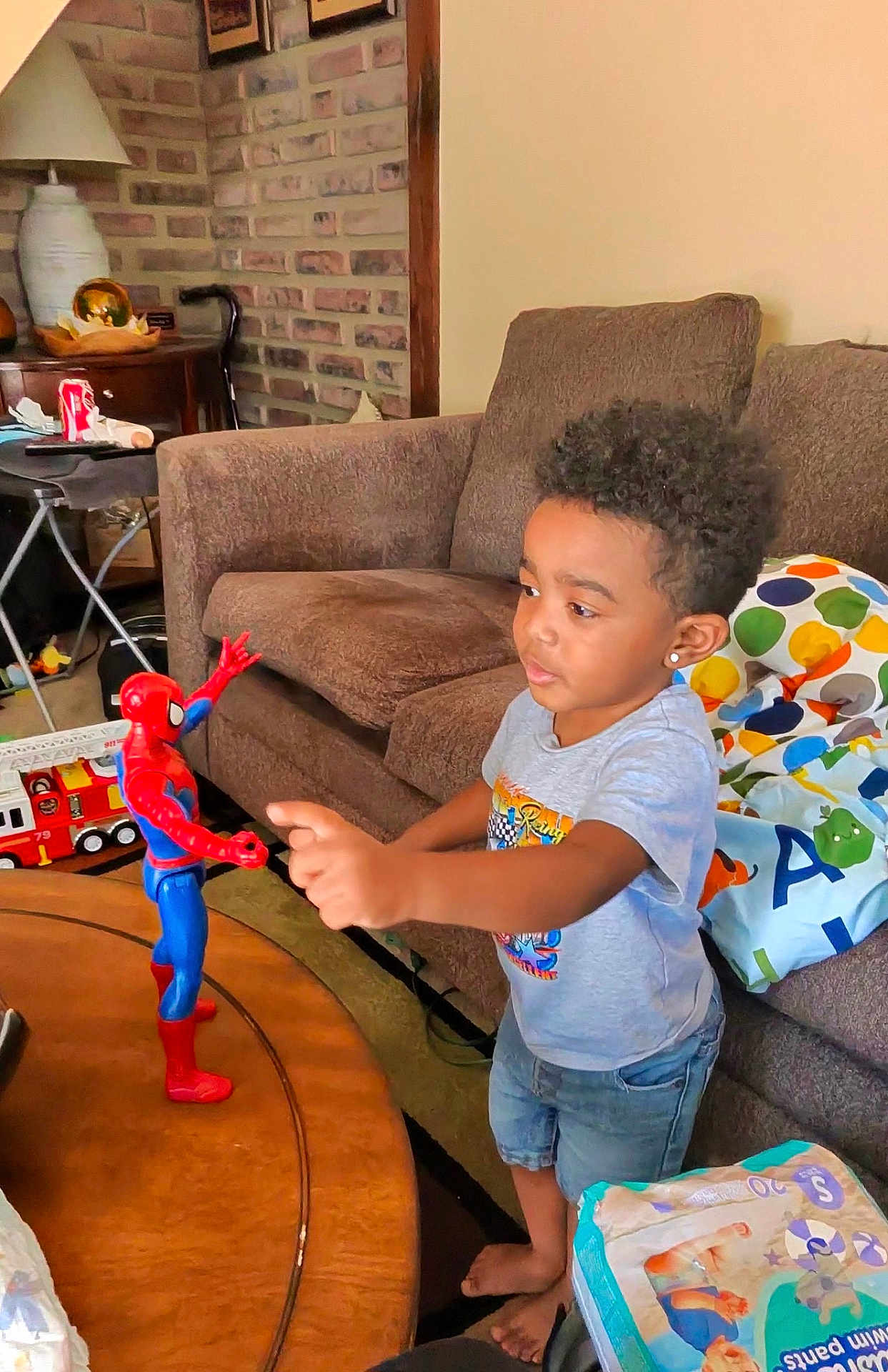 Zander is registered to the contest to win money with this photo: child, boy, action_figure, spider_man, toy, table, living_room, couch, pillow, brick_wall, lamp, diaper_pack, barefoot, curly_hair, casual_clothing, indoor, play, furniture, home, interaction