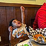 Zander is registered to the contest to win money with this photo: child, boy, food, table, restaurant, fried_food, french_fries, tshirt, happy, raising_arm, wooden_wall, spoon, book, checkered_paper, indoor, sitting, person, curly_hair, meal, casual_clothing