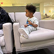 Zander joined the competition — help win amazing prizes! child, toddler, sitting, cookie, white_shoes, casual_clothing, armchair, indoor, waiting_area, modern_furniture, curly_hair, focused, neutral_colors, carpet, people_in_background, light_ceiling, person, holding_food, relaxed, side_profile