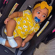 Zaniah is registered to the contest to win money with this photo: baby, child, car_seat, pacifier, yellow_clothing, headband, socks, indoor, floor, curly_hair, infant, cute, sitting, comfort, child_seat, black_car_seat, yellow, floral_pattern, young_child, portrait