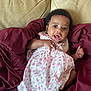 baby, infant, child, dress, floral_pattern, blanket, couch, sofa, maroon, beige, indoor, person, smiling, happy, cute, portrait, skin, hand, foot, sitting