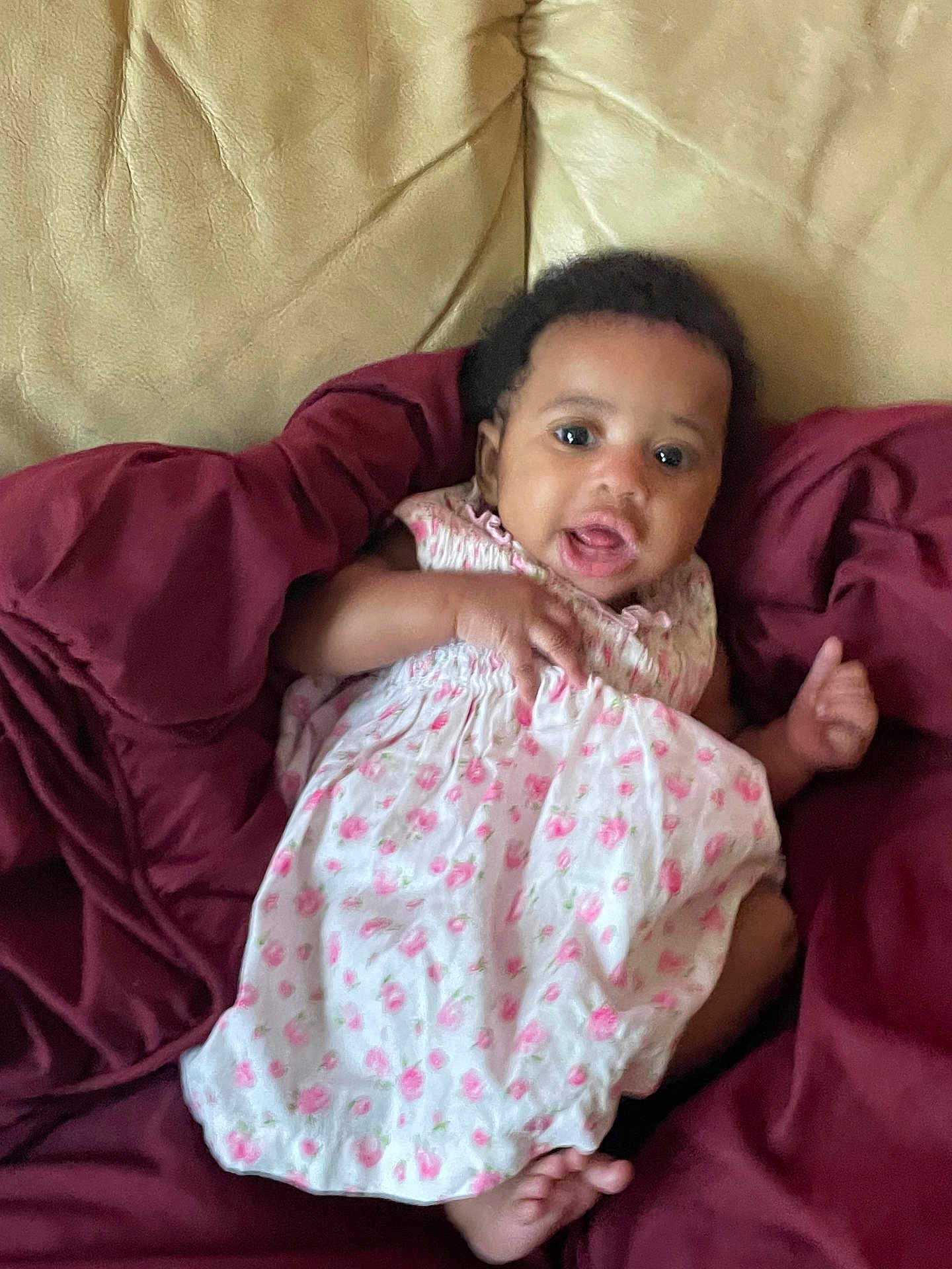Zyionnie is registered to the contest to win money with this photo: baby, infant, child, dress, floral_pattern, blanket, couch, sofa, maroon, beige, indoor, person, smiling, happy, cute, portrait, skin, hand, foot, sitting