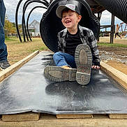 Malakai is registered to the contest to win money with this photo: child, boy, slide, playground, outdoor, casual_clothing, cap, jeans, plaid_shirt, smiling, happy, grass, wooden_structure, tube_slide, footwear, person, daylight, cloudy_sky, recreation, fun