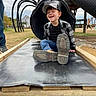 child, boy, slide, playground, outdoor, casual_clothing, cap, jeans, plaid_shirt, smiling, happy, grass, wooden_structure, tube_slide, footwear, person, daylight, cloudy_sky, recreation, fun