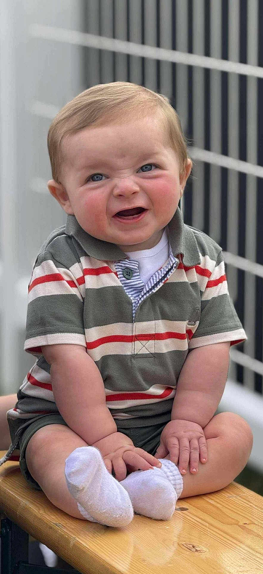 Andrew is registered to the contest to win money with this photo: baby, child, infant, smiling, sitting, striped_shirt, polo_shirt, white_socks, wooden_bench, outdoor, fence, cute, face, hands, legs, blue_eyes, short_hair, skin, expression, portrait