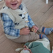Léon a rejoint le concours — aidez-le/la à gagner de superbes lots ! toddler, child, smiling, indoor, wooden_floor, fleece_vest, plaid_shirt, striped_socks, toy, wooden_toy, plush_toy, woven_rug, happy, sitting, cute, young_child, home, person, casual_clothing, playtime