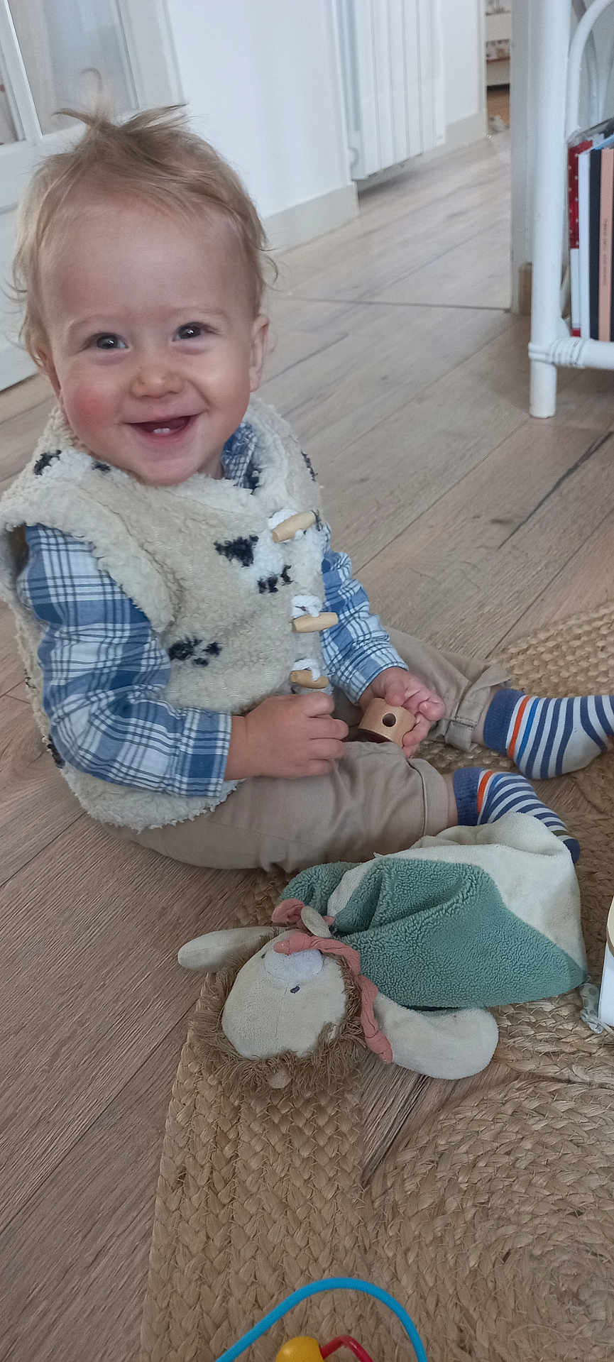Léon a rejoint le concours — aidez-le/la à gagner de superbes lots ! toddler, child, smiling, indoor, wooden_floor, fleece_vest, plaid_shirt, striped_socks, toy, wooden_toy, plush_toy, woven_rug, happy, sitting, cute, young_child, home, person, casual_clothing, playtime